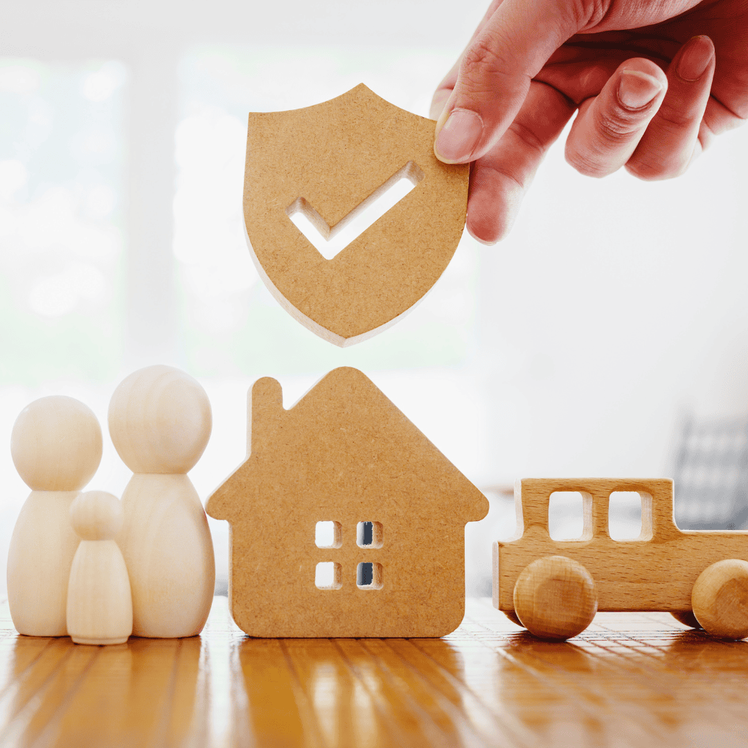 A hand places a wooden shield with a checkmark above a miniature wooden house, family figures, and a car, symbolizing home security and protection.