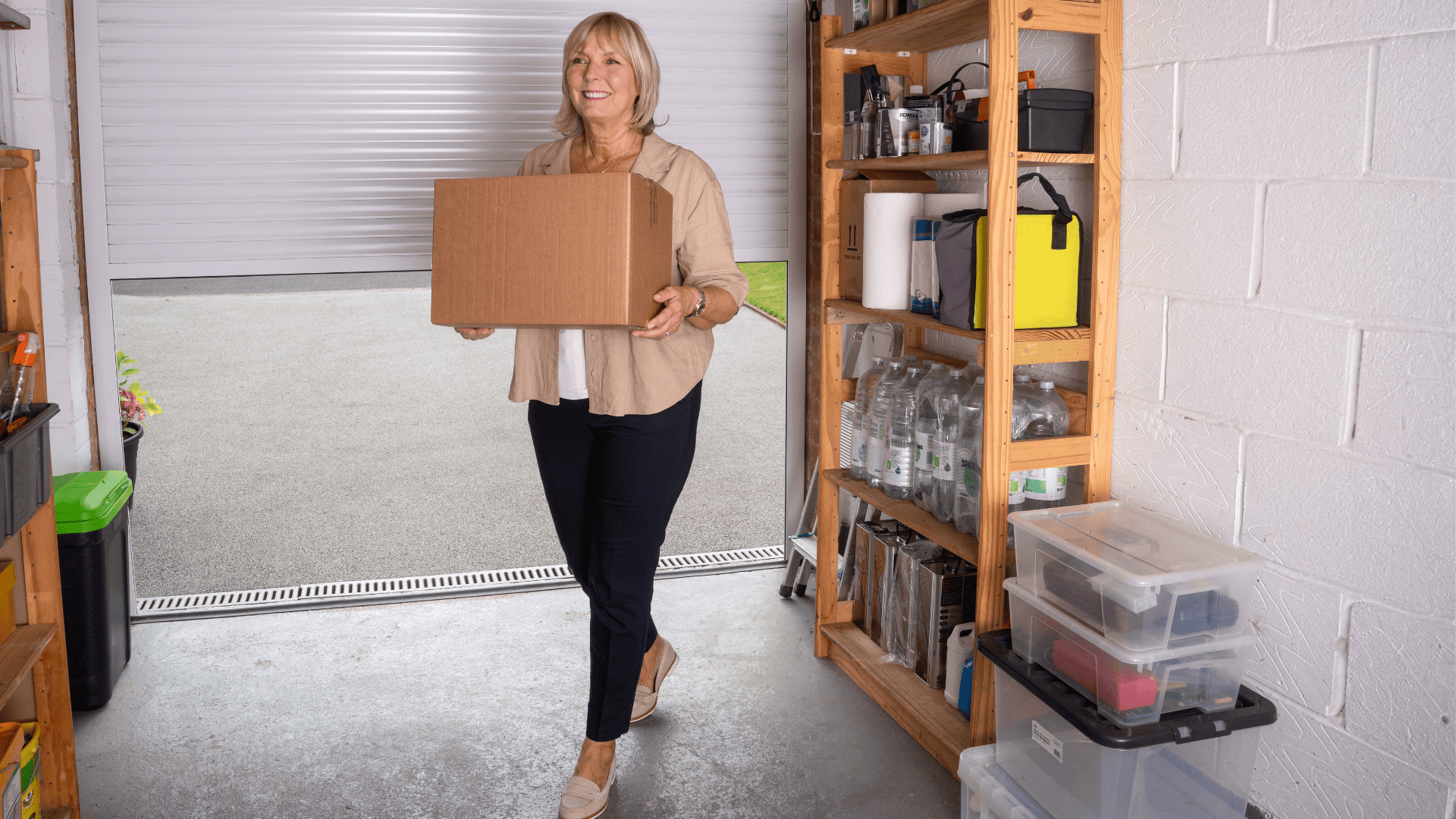 A smiling woman carrying a cardboard box inside a well-organized garage with shelves of storage containers and bottled water. The garage door is open.