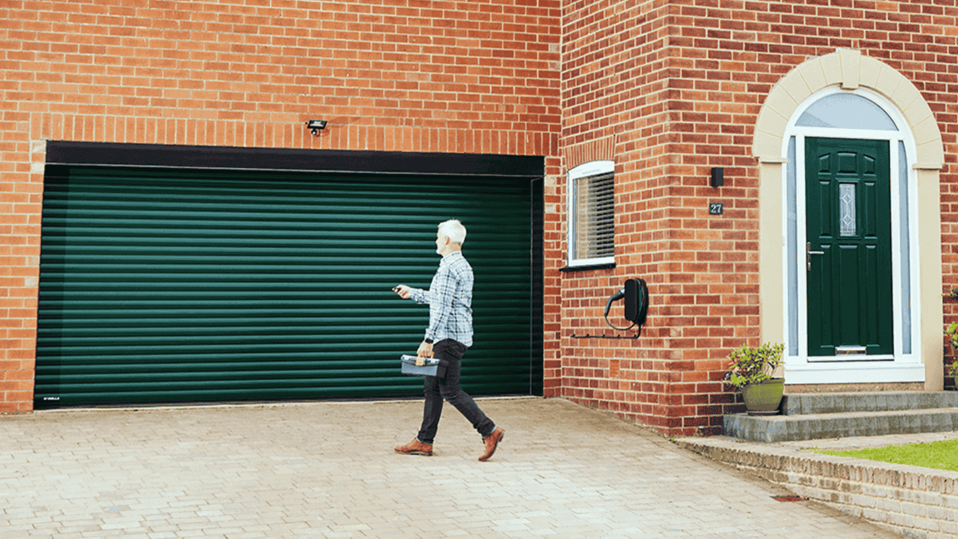 A man in a checkered shirt walks towards a house with a dark green roller garage door, using a remote control. The house has a matching green front door.