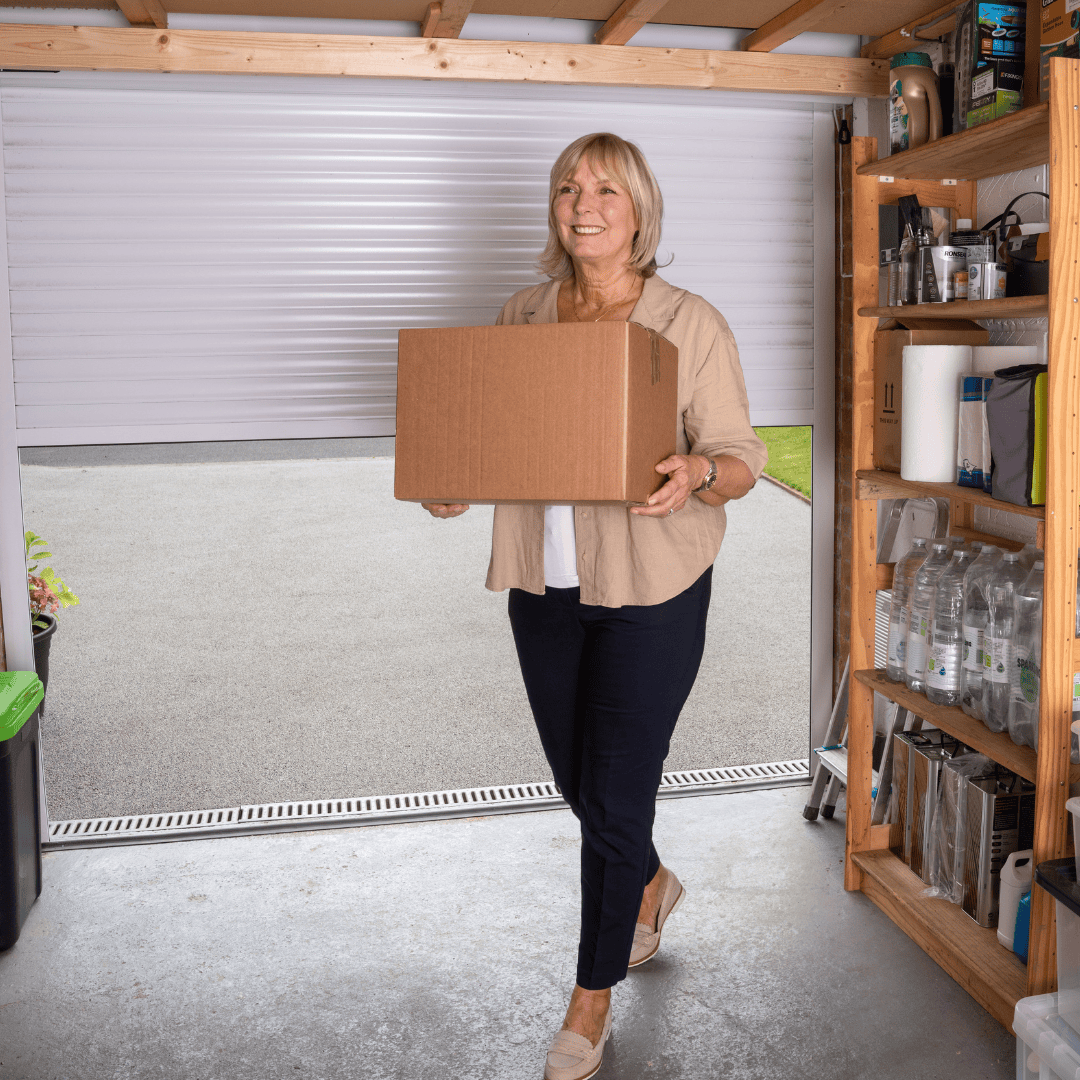 A smiling woman carrying a cardboard box inside a well-organized garage with shelves of storage containers and bottled water. The garage door is open.