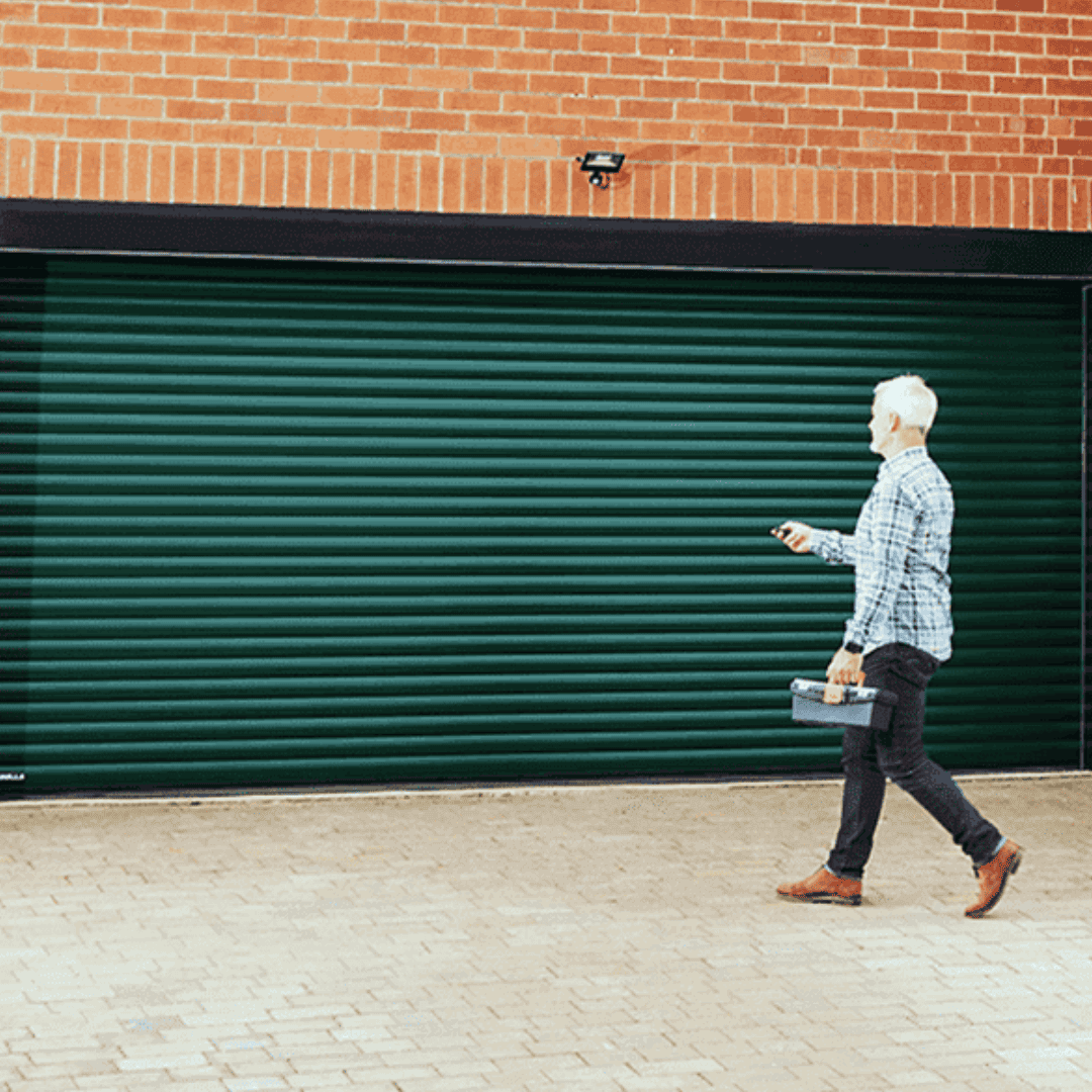 A man in a checkered shirt walks towards a house with a dark green roller garage door, using a remote control. The house has a matching green front door.