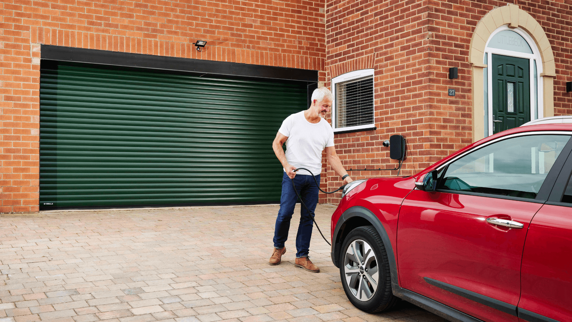 Homeowner charging a red electric car on a driveway with a green insulated roller garage door and wall-mounted EV charger in the UK