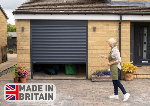 A woman opening a grey electric roller garage door at a suburban home, revealing garden equipment inside, with a Made in Britain logo