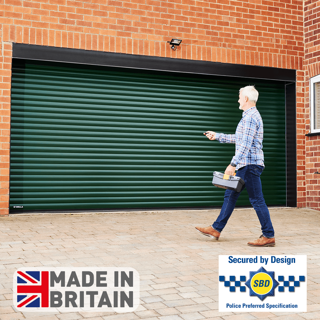 A man in a checkered shirt walks towards a house with a dark green Titan roller garage door, using a remote control. The house has a matching green front door.